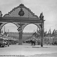 Feria de 1933. La sobria portada luce el escudo de armas de la II República española y queda rematada a ambos lados con la alegoría de la República. Circulan bajo ella automóviles y caballistas.