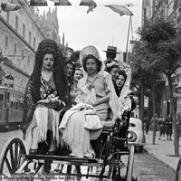 Ataviadas con mantones y mantillas se dirigen al Real en un coche de colleras por la avenida de José Antonio (hoy Constitución). A la izquierda, un tranvía junto a la Punta del Diamante y a la derecha un dispensador de gasolina. Década de 1940.