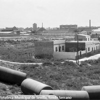 11. Barriada de El Turruñuelo, 1964. Las calles del Turruñuelo fueron adoquinadas en 1940 y rotuladas en 1950. Pero no fue hasta mediados de los años sesenta cuando se acometieron obras de saneamiento e infraestructura. En el horizonte, la fábrica de espejos y cristales de Pueyo y las chimeneas de los tejares que compiten en altura con la Torre de Los Remedios.  ©ICAS-SAHP. Fototeca Municipal de Sevilla, fondo Serrano