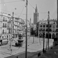 Aún perduran algunos de los soportales de la línea de casas que desde mediados del XIX vienen desapareciendo. Un suntuoso quiosco de bebidas resalta en la imagen, cuya función, desde 1895, era la de proporcionar agua fresca en zonas públicas. Este uso se alternaba con la venta de billetes del servicio de tranvías. La plaza aparece preparada para la conmemoración de la fiesta del Corpus a falta de colocar las velas, gallardetes y colgaduras vegetales. [1916-1917].  © ICAS-SAHP, Fototeca Municipal de Sevilla, fondo Serrano